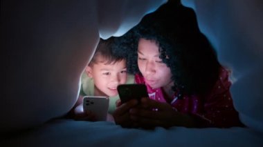 Afrika kökenli Amerikalı genç bir annenin gecenin karanlığında küçük oğluna iyi geceler masalı okuması ve akıllı telefonların aydınlattığı yüzler. Aile sevgisi eğitim konsepti 4K