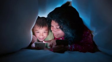 Küçük çocuk rahat bir yatakta battaniyenin altında akıllı telefonla oynuyor. Afrika kökenli Amerikalı genç bir annenin karanlık bir gecede battaniyenin altında oğluna iyi geceler hikâyesi okuması ve yüzlerinin ekranlarla aydınlatılması.