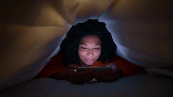 スマートフォンで色スクロールフィードの女性 巻き毛読書本を持つ若いアフリカ系アメリカ人女性のスローモーションや毛布の下で暗闇の中でソーシャルメディアをスクロールします 自由時間教育の概念 — ストック動画