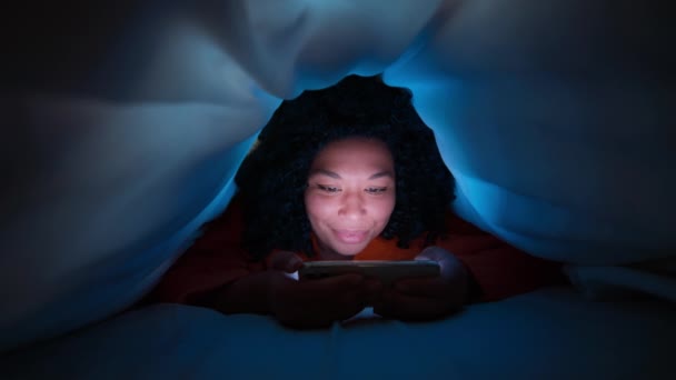 巻き毛読書本を持つ若いアフリカ系アメリカ人女性のスローモーションや毛布の下で暗闇の中でソーシャルメディアをスクロールします 自由な時間教育の概念 スマートフォンでカラースクロールフィードの女性 — ストック動画