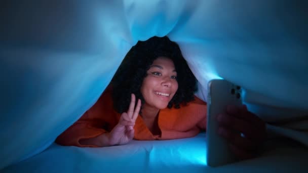 毛布の下に幸せなアフリカ系アメリカ人女性 アフロヘアの女性がSnsで写真を撮ったり スマートフォンのビデオ会議を使って友人と話をしたりして 夜にベッドの上で寝る — ストック動画
