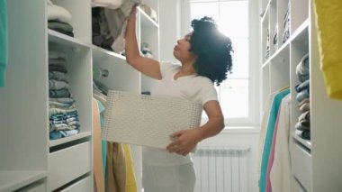 Siyah kıvırcık saçlı, kirli çamaşırlarını çıkartıp çamaşır sepetine koyan ve çamaşır makinesine giden genç bir kadın. Hizmetçi evde çalışıyor, yavaş çekim bahar temizliği konsepti 4K