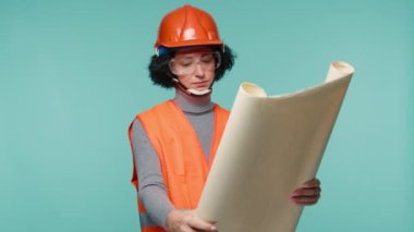 Stüdyodaki izole edilmiş mavi arka plan üzerine mimari tasarım üzerinde çalışan çekici, ciddi ve sorumlu mimarın ön görüşü. Turuncu miğfer ve yelek giymiş 4K tasarımcı görüntüleri.