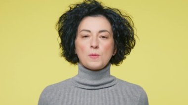 40 'lı yaşlarda gri kazaklı, gülümseyen olgun bir kadın kameraya bakıyor sakız çiğniyor balon şişiriyor sarı stüdyo arka planında izole edilmiş topu patlatıyor. Halk yaşam tarzı konsepti