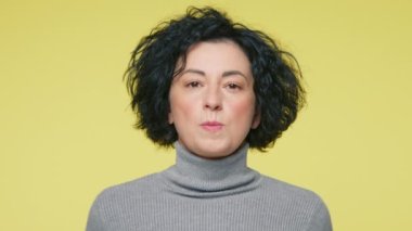 Gülümseyen olgun bir kadın, sarı stüdyo arka planında izole edilmiş bir şekilde taşaklarını patlatır. 40 'lı yaşlarda gri kazak giyen, kameraya bakıp balon sakızı çiğneyen insanlar.