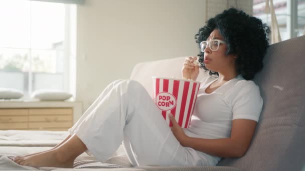 害怕的非洲裔美国女性在沙发上看电影 在家里吃爆米花 被恐怖片情节吓到的女性 在家里4K天 迷人的女人拿着爆米花篮子坐在沙发上 — 图库视频影像