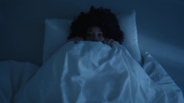 アフリカ系アメリカ人の女性は 暗い夜に毛布の下に隠れて ベッドの上で幽霊を怖がって怖がらせた ベッドの上に横たわる色の女性の上のビューと自宅で怖いものを見ないように毛布で顔をカバー4K — ストック動画