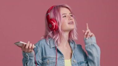 Yavaş çekim sümüklü, modaya uygun pastel pembe saçlı, kırmızı kulaklıklı, akıllı telefonlu, kapalı mekanda dans eden, yalnız başına eğlenen hippi kız. Stüdyoda pembe arka planda dans eden mutlu genç kadın.