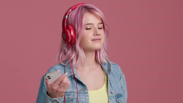 快乐的年轻女子在粉红背景的工作室里跳舞 身披红色耳机 留着时髦的粉红头发 手持智能手机 手舞足蹈在室内的慢动作瘦小嬉皮士女孩 一个人玩4K — 图库视频影像