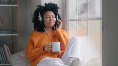 Young relaxed African American woman using headphone while slowly dancing at window. Black woman using app on smartphone at home while listening music. Casual girl with afro hairstyle drinking hot tea