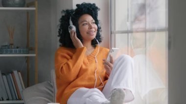 Smiling young mixed race student girl listening music in headphones with phone moving slowly indoors on rainy cold day, feeling free. Happy cool African American woman dancing while sitting at window