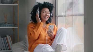 Smiling young mixed race lady hipster listening music indoors on rainy cold day, feeling free. Happy cool African American woman in headphones with phone moving slowly, dancing while sitting at window