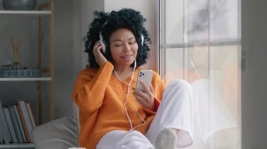 Happy cool African American woman in headphones with phone moving slowly, dancing while sitting at window. Smiling young mixed race lady hipster listening music indoors on rainy cold day, feeling free