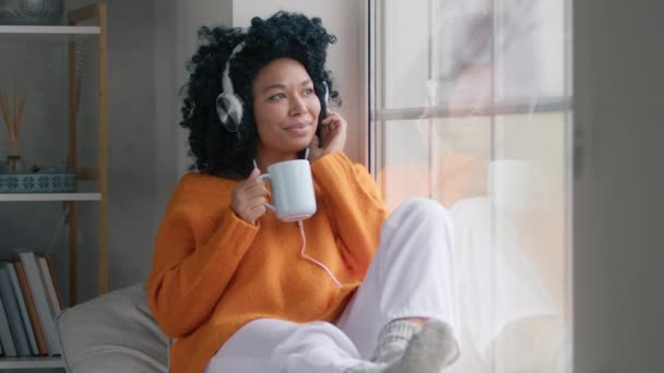 アフロヘアスタイルのカジュアルな女の子は熱いお茶を飲む 若いリラックスしたアフリカ系アメリカ人女性がゆっくりと窓で踊りながらヘッドフォンを使用しています 音楽を聴きながら自宅でスマホアプリを使う黒人女性 — ストック動画