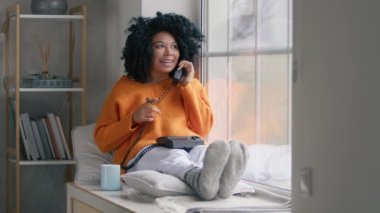Indoor shot in stylish living room on cold rainy day. Good looking dark haired curly woman in orange sweater sitting at large window with handset phone talking with boyfriend with flirting expressing