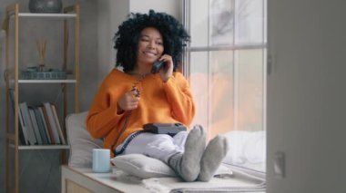 Good looking dark haired curly woman in orange sweater sitting at large window with handset phone talking with boyfriend with flirting expressing. Indoor shot in stylish living room on cold rainy day