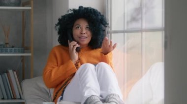 Portrait of african american woman in orange sweater with handset phone actively discussing life situation with friend sitting at large window. Girl with afro hairstyle talking sitting at large window