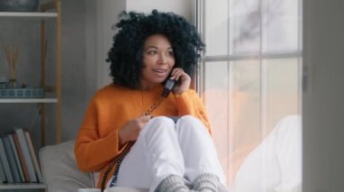 Portrait of Good looking dark hair curly woman in orange sweater sitting at large window with handset phone talking with boyfriend with flirting expressing. Indoor shot in stylish room on rainy day