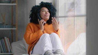 Portrait of african american woman in orange sweater with handset phone spreading rumors tittle-tattle with girlfriend sitting at window. Girl with afro hairstyle gossiping on rainy spring weekend day
