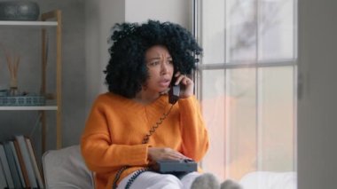 Portrait of african american woman in orange sweater sitting at window with handset phone swear at boyfriend over the phone. Upset girl with afro hairstyle got angry, yelling into the phone on rainy