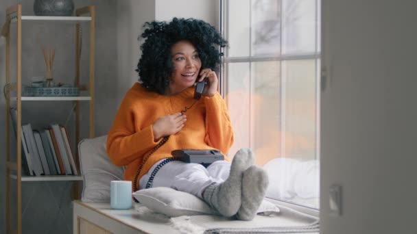 携帯電話でオレンジ色のセーターを着たアフリカ系アメリカ人女性を笑いながら 大きな窓に座って彼氏といちゃつく 屋内ショット女の子とともにAfro毛深いです話している間に座って大窓 — ストック動画