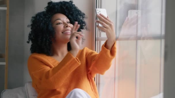 中暗い髪の女の子は ウィンドウで携帯電話のカメラで写真を撮る Vの勝利の兆候を示す 空気のキスを送信します アフリカ系アメリカ人ソーシャルメディアブロガー出身 スマートフォンを使う女 — ストック動画