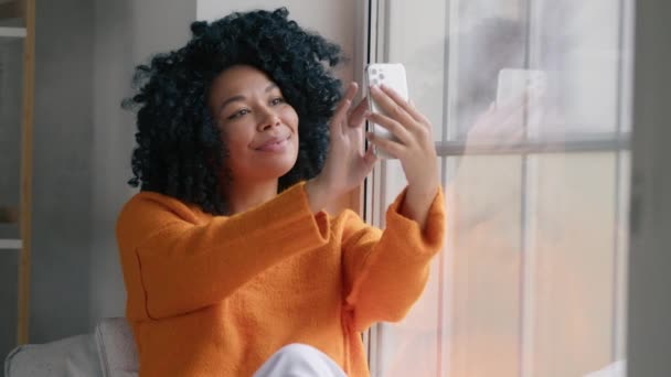 年轻女人在家里用智能手机 非洲裔美国女孩坐在窗前看着手机摄像头 社交媒体博客作者从家里开始工作 连接移动应用技术生活方式概念 — 图库视频影像