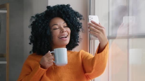 自宅から働くアフリカ系アメリカ人のソーシャルメディアブロガー 接続モバイルアプリ技術ライフスタイルの概念 家でスマホを使っている若い女性 窓に座っている携帯電話のカメラを見ている女の子 — ストック動画