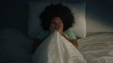 Adult african american woman scared by noise in home or hotel room. Girl with afro hairstyle infantility hiding under blanket, looking around scared to see the monster or ghost, slow motion nightmare