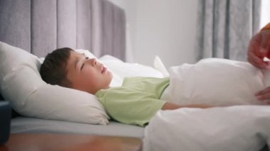 Mother caring for sick mixed race boy lying in bed with flu and cough, measuring temperature using hand. Little preschool boy at home with fever. African american mom giving health care slow motion
