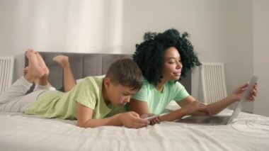 Mom using laptop and boy holding smartphone enjoying online fun video game play. Young generation internet technology addiction.African American woman with her preschool son lying on bed using gadgets