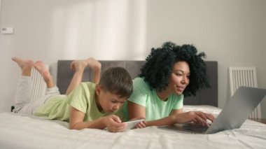 Young generation internet technology addiction.African American woman with her preschool son lying on bed using gadgets. Mom using laptop and boy holding smartphone enjoying online fun video game play