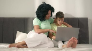Black family looking at laptop in home interior. Puzzled preschool boy solving math exercise with smiling mother. African American woman and child make video call sitting on bed in bedroom sunny day