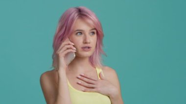 Closeup sweet hipster girl speaking mobile phone indoors. Portrait of happy positive woman with pink hair using smartphone. Cheerful female person chatting on cellphone on blue wall studio background