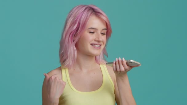 若い女性の笑顔20代のピンクの髪は スピーカーフォンで音声メッセージを記録 バーチャルアシスタントアプリを使用している学生は スタジオのバックグラウンドでスマートフォンにリマインダーを設定します デジタルAiモバイル支援技術ショット — ストック動画