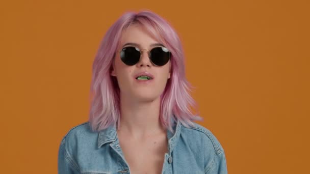 戴现代太阳镜的女人慢吞吞地咀嚼口香糖 穿着蓝色斜纹棉布的嬉皮士粉色头发的性感小女孩站在橙色背景下吹泡糖泡 — 图库视频影像