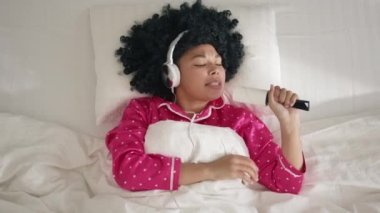 Slow motion of happy young African American woman with afro hair in headphones dancing and singing in clothes roller having fun lying in comfy bed on sunny morning. People, lifestyle and joy concept.