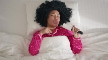 Happy cute African American girl 20s having fun in bed on sunny morning. Funny mixed race woman dancing while lying in comfortable bed on white linen pillow, singing in sticky roller like microphone