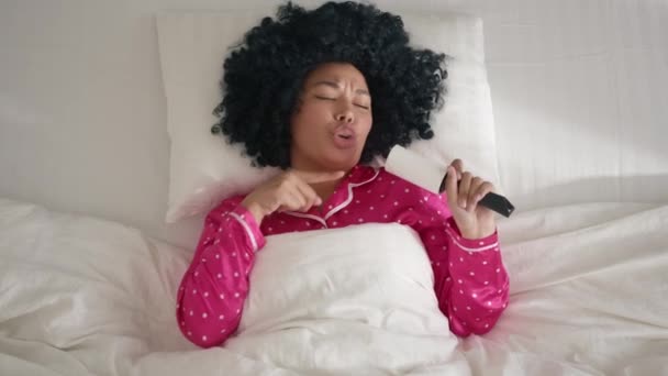 幸せなかわいいアフリカ系アメリカ人の女の子20代は晴れた日の朝にベッドで楽しんでいます 白いリネンの枕の上に快適なベッドに横たわっている間面白い混合レースの女性は マイクのような粘着性のローラーで歌って踊る — ストック動画