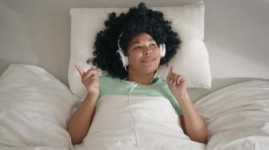 Happy cool African American woman of color wearing headphones dancing alone lying in bed on sunny morning. Smiling young mixed race girl listening music in bedroom indoors, feeling free and funky 4K