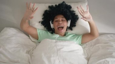 Happy cool African American woman of color wearing headphones dancing alone lying in bed on sunny morning looking in camera. Smiling young girl listening music in bedroom indoors feeling free funky 4K
