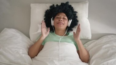 Top down view on Smiling afro hair girl listening music in bedroom indoors feeling free funky 4K. happy cool African American woman of color wearing headphones dancing alone lying in bed sunny morning