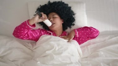 Funny cute woman of color in hot pink pajama lying in bed on sunny morning, having fun celebrating successful lifestyle, dancing enjoying music on weekend. Happy African American woman singing at home