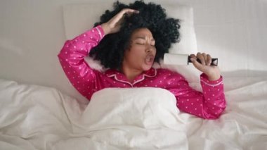 Happy African American woman singing at home. Funny cute woman of color in hot pink pajama lying in bed on sunny morning, having fun celebrating successful lifestyle, dancing enjoying music on weekend