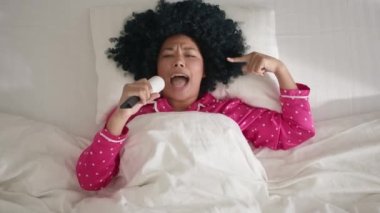 Close-up view from above woman of color in pink pajama, beautiful cheerful girl with afro hairstyle, happy woman sings in sticky clothes roller having fun singing song dances to music, bedroom morning
