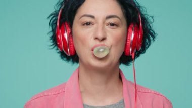 Close up portrait of positive mature caucasian woman with short dark curly hair listening to music with red headphones and blowing bubble from the bubblegum, laughing in green wall studio background