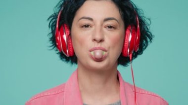 Close up portrait of positive mature caucasian woman blowing bubble of chewing gum. 40s model with short dark curly hair listening to music with red headphones and laughing in green studio background