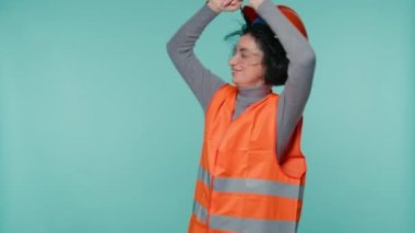 Smiling portrait of mature caucasian female industrial engineer in orange hard hat, safety vest happily dancing on blue green studio background. Inspector or Supervisor Industrial Construction 4K