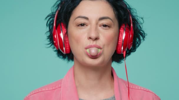 正成熟的高加索女人吹制口香糖泡泡的特写 40年代的模特 黑发短发 用红色耳机听音乐 在绿色工作室的背景下大笑 — 图库视频影像