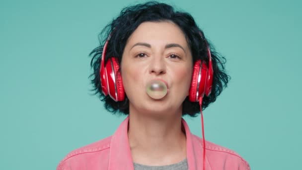 赤いヘッドフォンを持つ正成熟した白人女性の肖像画を閉じ 緑のスタジオの背景で笑って ガムの泡を吹いて 音楽を聴く短い暗い巻き毛の40年代モデル — ストック動画